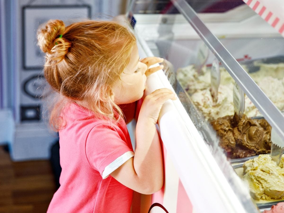 A young girl in a pink shirt presses against an ice cream display case, gazing intently at several flavors as she tries to choose.