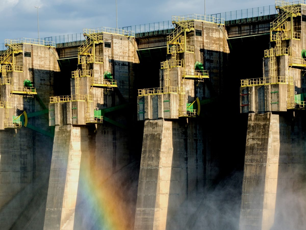 Hydroelectric dam with yellow railings and visible mist, featuring a faint rainbow at the base of the structure