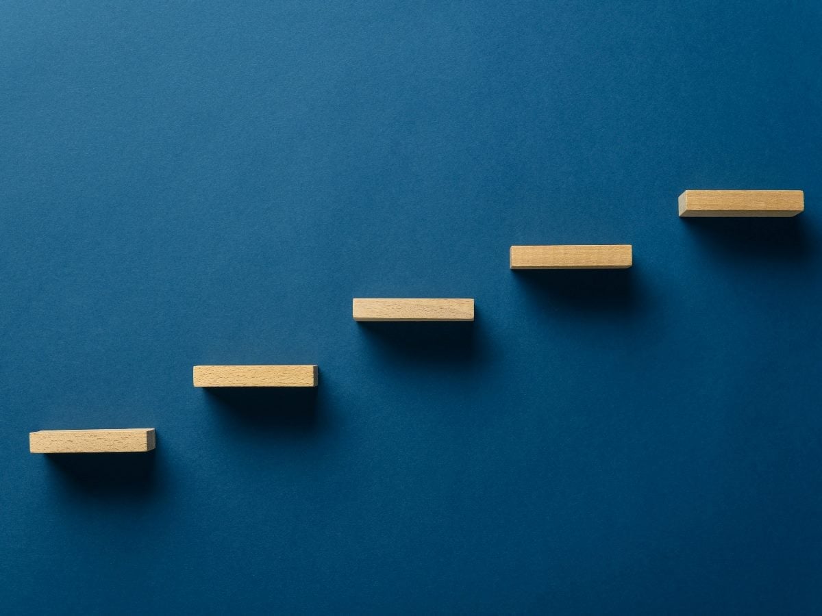 Five wooden blocks arranged as ascending steps against a blue background, symbolizing a structured, step-by-step progression or framework.