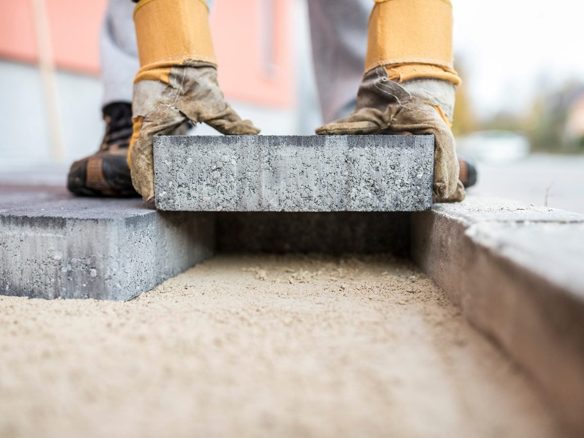 Close-up photograph of a worker wearing protective gloves carefully placing a concrete paver onto a prepared sand base. The image emphasizes the foundation layer beneath the paving stones, illustrating the importance of proper groundwork and structural support before installation.