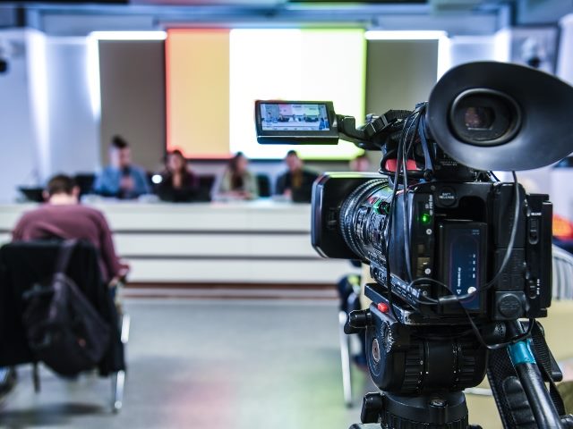 A television camera pointed toward a table with people sitting at it in a press conference.