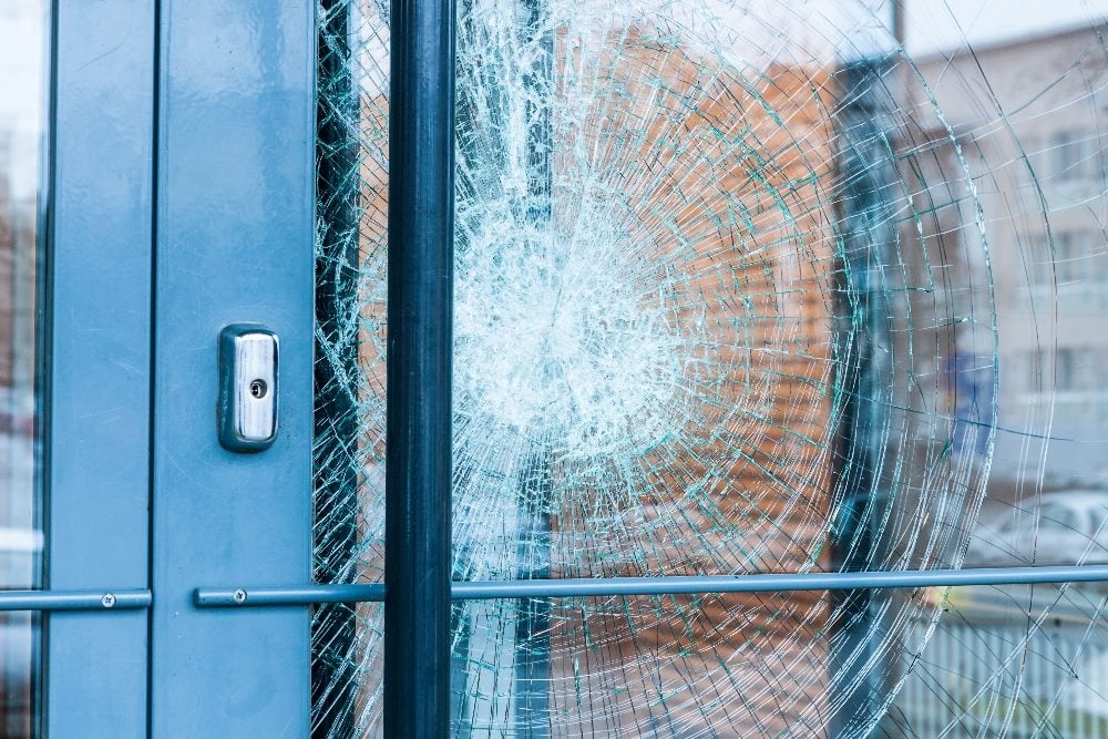 A close-up view of a glass door with a significant crack, radiating from a central impact point, creating a spiderweb-like pattern. The crack suggests a strong impact or force applied to the door. The door has a metal frame and a locking mechanism, and the background shows an urban setting with buildings slightly blurred in piece about the recent AT&T data breach.