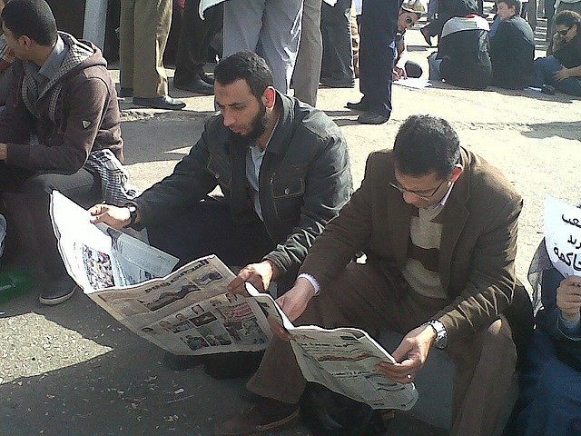 Two men sitting down outside reading newspapers.