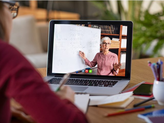a woman takes notes as she listens to a woman instructor in a video on her laptop.