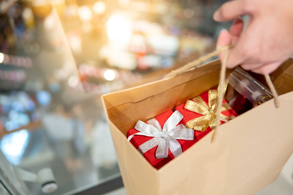 Closeup of a person's hand holding a brown craft paper shopping bag filled with beautifully colorfully wrapped and beribboned gifts with a sparkling background in piece about the upcoming holiday shopping season.