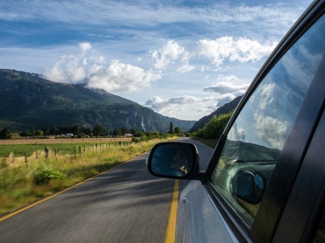 A car's side-view-mirror on a road in daytime with a mountain as a backdrop.