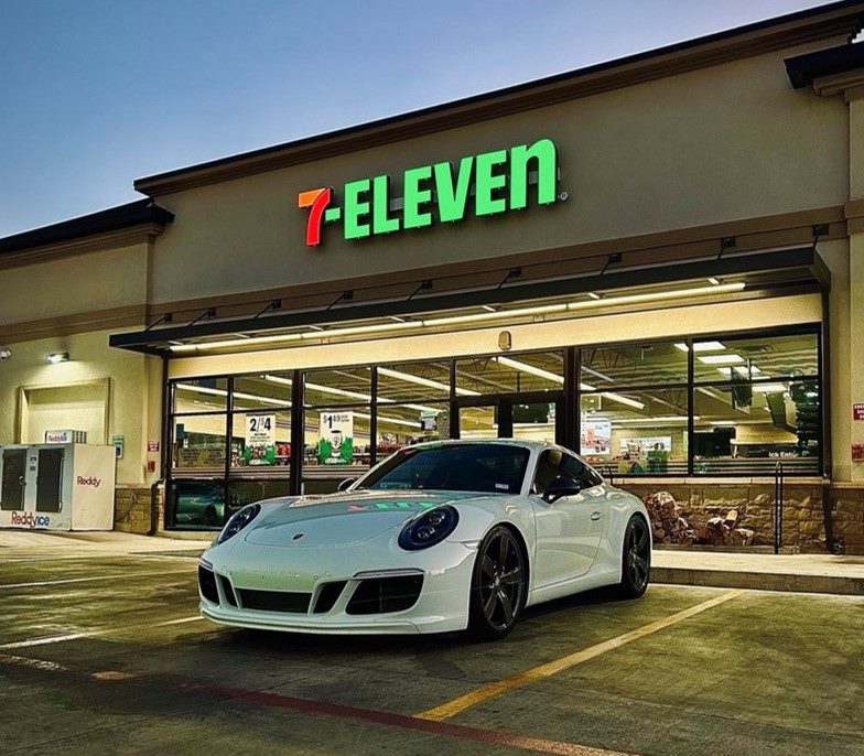 A sleek white Porsche sports car is parked in front of a 7-Eleven convenience store during twilight. The neon lights of the store illuminate the area, creating a contrast between the high-end car and the everyday retail setting.. This image suggests 7-Eleven's successful efforts at building strong customer communities..