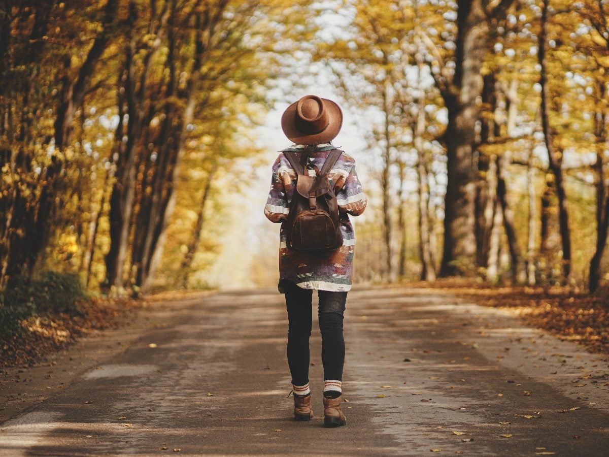 A person wearing a hat, backpack and fall clothing walks alone down a tree-lined road covered in autumn leaves.