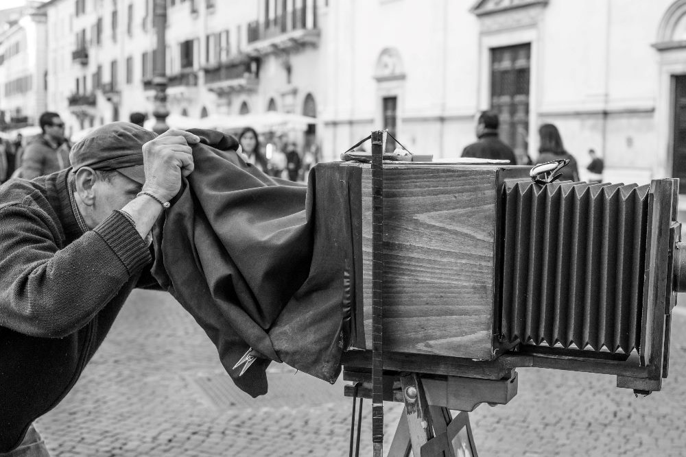 An elderly man wearing a cap is using an antique large-format camera in a busy outdoor setting. He is bent over, looking through the viewfinder while draped under a dark cloth for better visibility. The camera is mounted on a tripod, and the background shows a cobblestone street with people and historical buildings in a European city. The image is in black and white, emphasizing the vintage feel of the scene in piece about text-to-image generators.