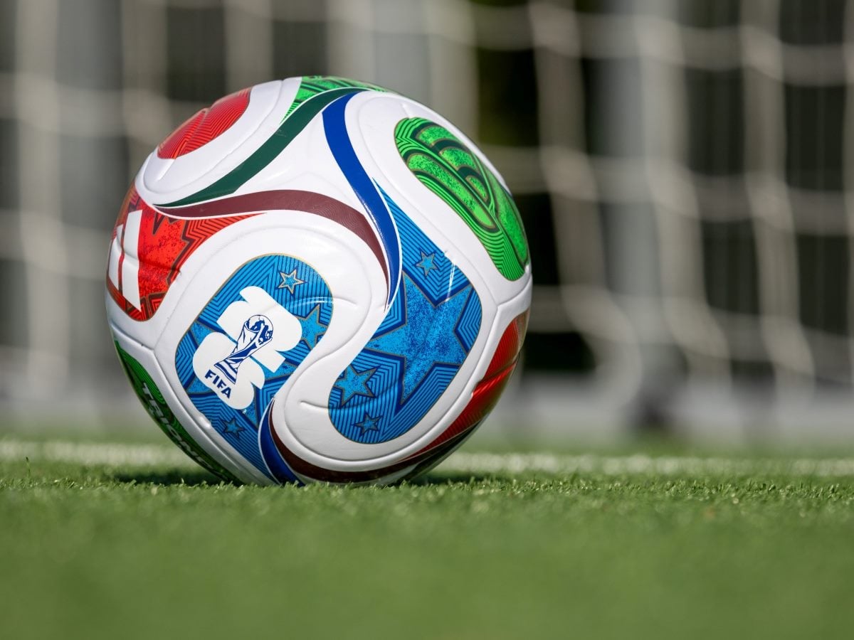 A close-up photograph of the official FIFA World Cup 2026 match ball resting on green turf in front of a soccer goal net. The ball's white surface features a colorful swirling design with red, green, and blue panels decorated with star patterns and thin outline graphics, along with the FIFA logo and a trophy icon printed near the center. The soccer goal's net and white post are visible blurred in the background, indicating the photo was taken on the pitch near the goal line.