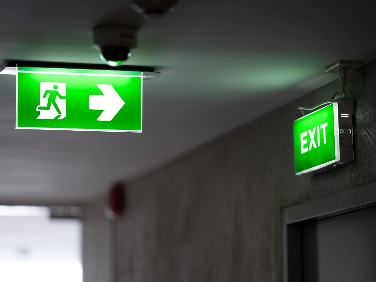 A pair of illuminated green exit signs mounted on a dim, low-lit hallway ceiling and wall, one showing a running figure and directional arrow, the other reading "EXIT" in bold white letters, with a security camera and smoke detector visible nearby against bare concrete and plaster surfaces.