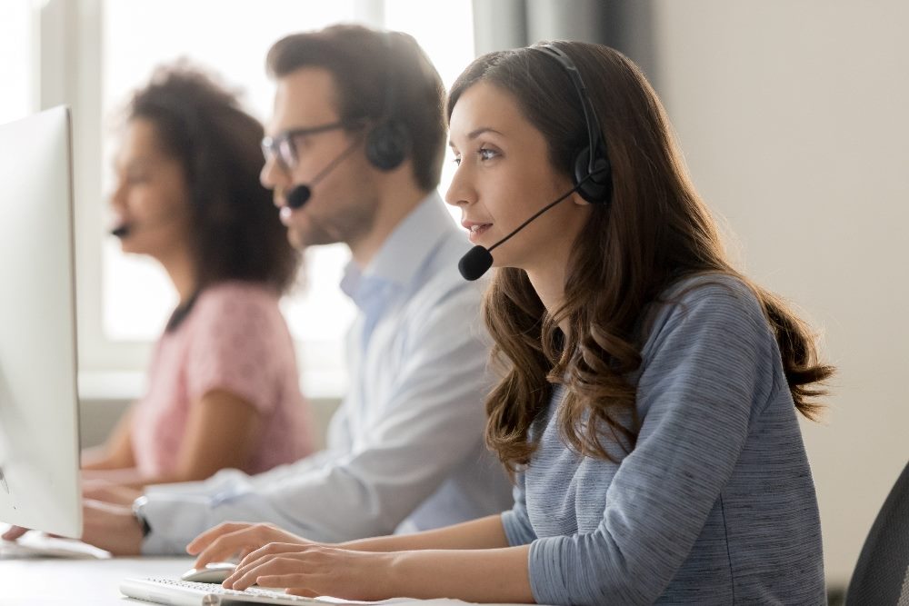 Three contact center agents sit at tables in front of computers and wearing headsets while consulting with callers, suggesting the changes occurring as AI in contact centers becomes more prevalent.