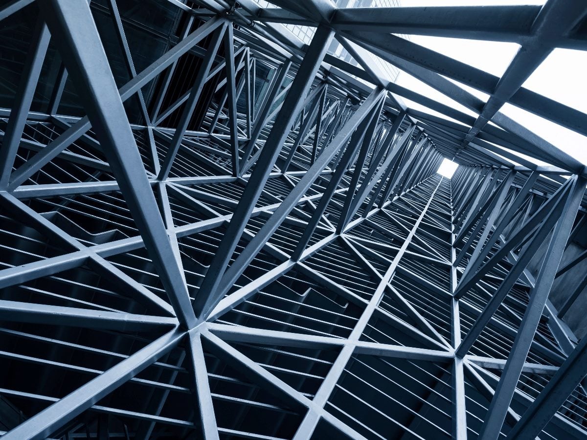 Low-angle view of a modern building’s steel framework and lattice structure, with intersecting beams and geometric lines rising upward toward a bright sky, emphasizing architectural scale and structural design.