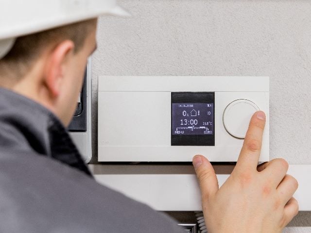 An engineer wearing a white construction hat and gray shirt adjusts a thermostat for automated heating system in piece about business transformation and managing expectations of CIOs. 