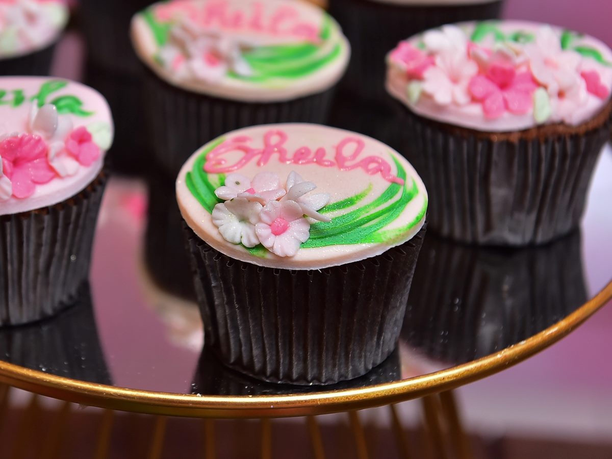 Close-up of decorated cupcakes with pink and white floral icing and green accents; one cupcake in focus has the name “Sheela” piped in pink frosting on top.