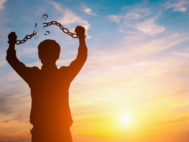 A silhouetted  man in front of a sunset. His hands are over his head as he breaks the chains