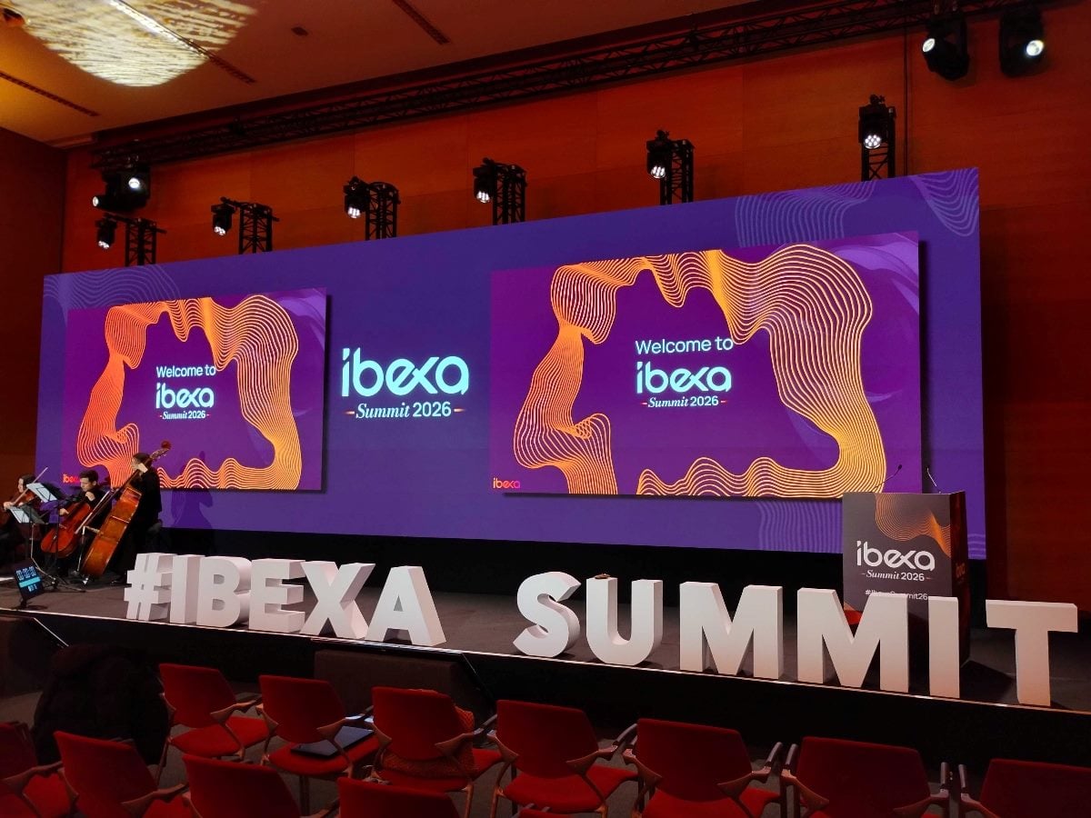 Wide view of the Ibexa Summit 2026 main stage, featuring large purple-and-orange digital screens reading “Welcome to ibexa Summit 2026,” #IBEXA SUMMIT lettering across the stage front, and an empty audience seating area in a modern conference hall.