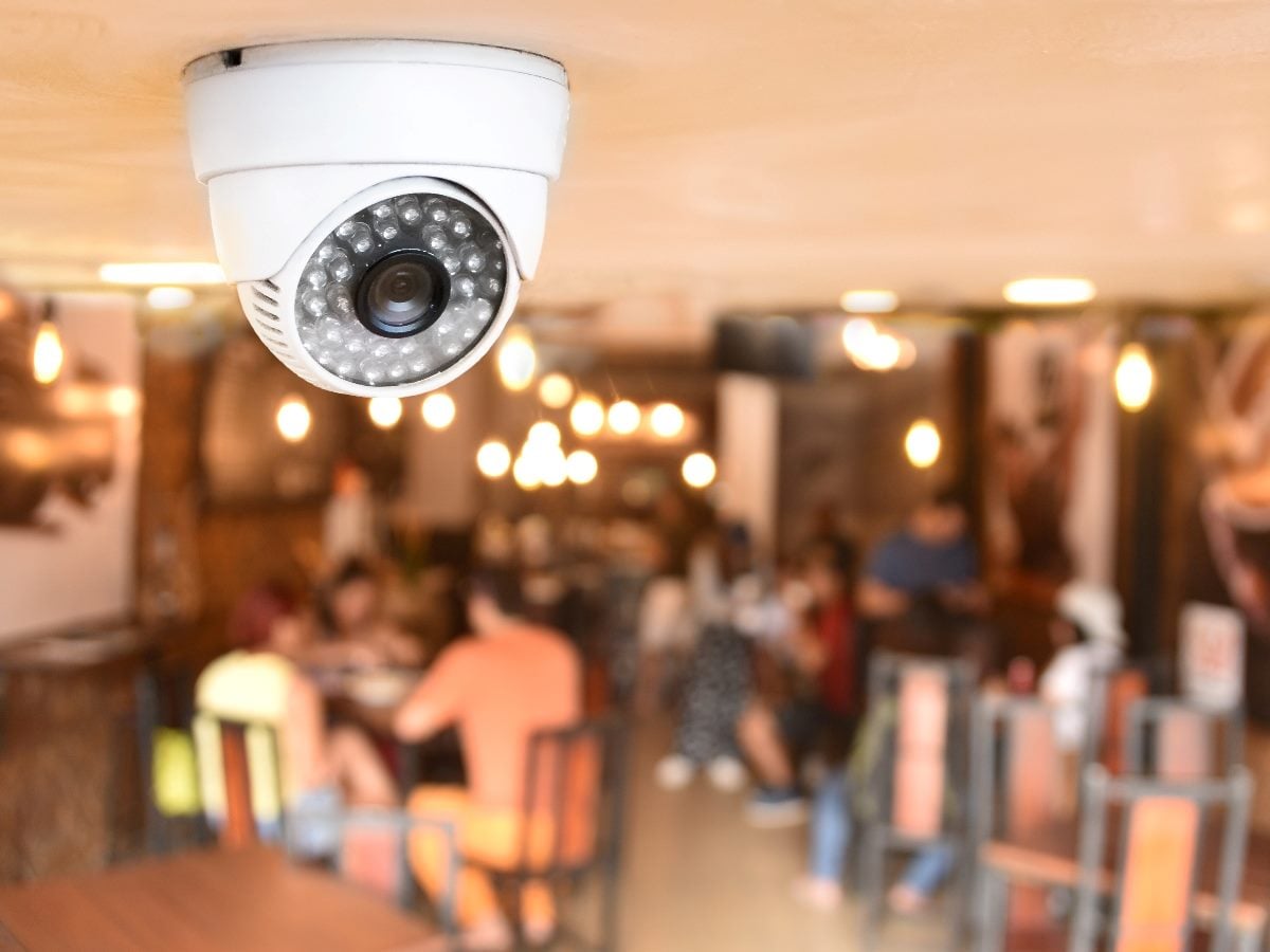 Dome-style security camera mounted on a ceiling in the foreground, sharply in focus, overlooking a blurred café interior with customers seated at tables under warm lighting.