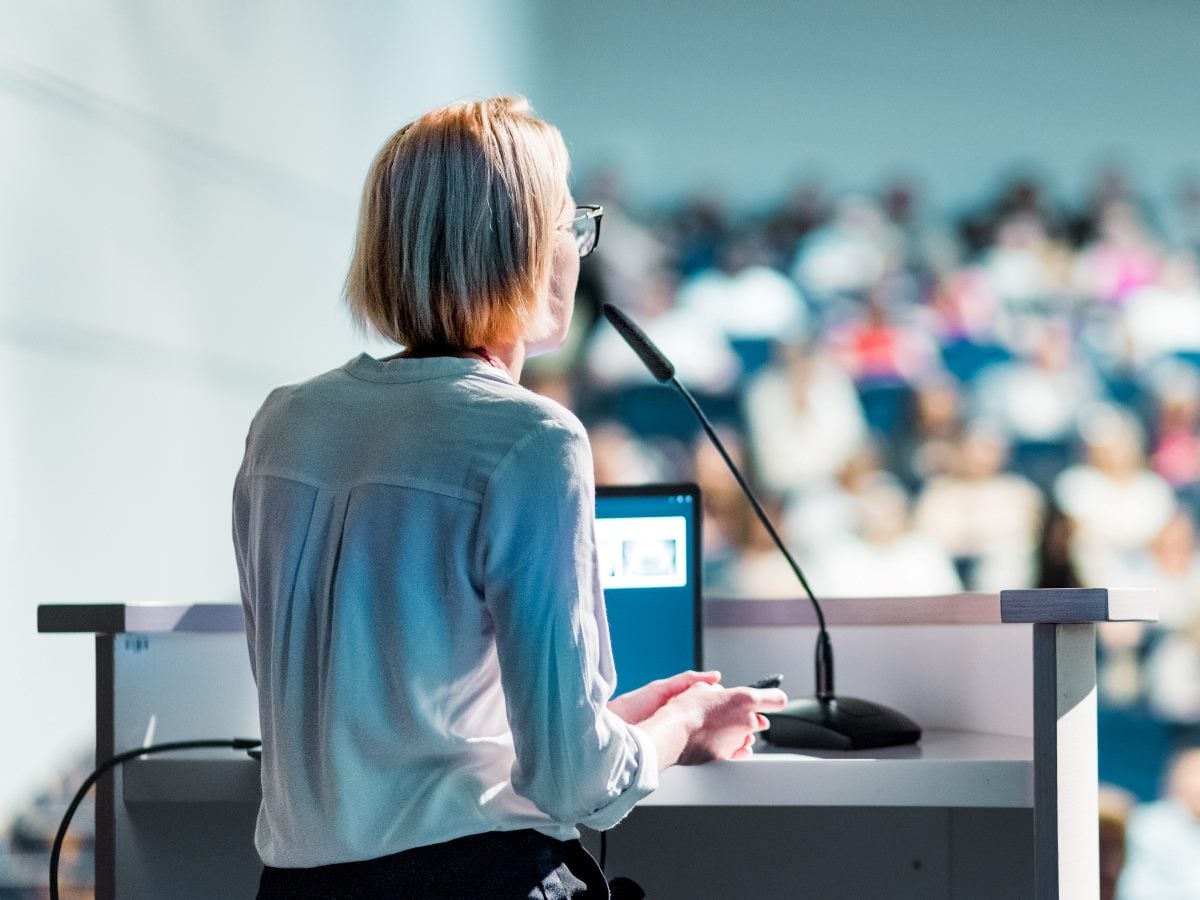 A business professional speaks at a podium with a microphone, facing a large, blurred audience in a conference or keynote setting.