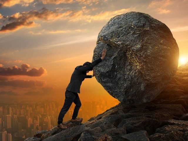 A man pushing large stone up to hill against a sunset in the background symbolizing the need to improve the customer effort score. 