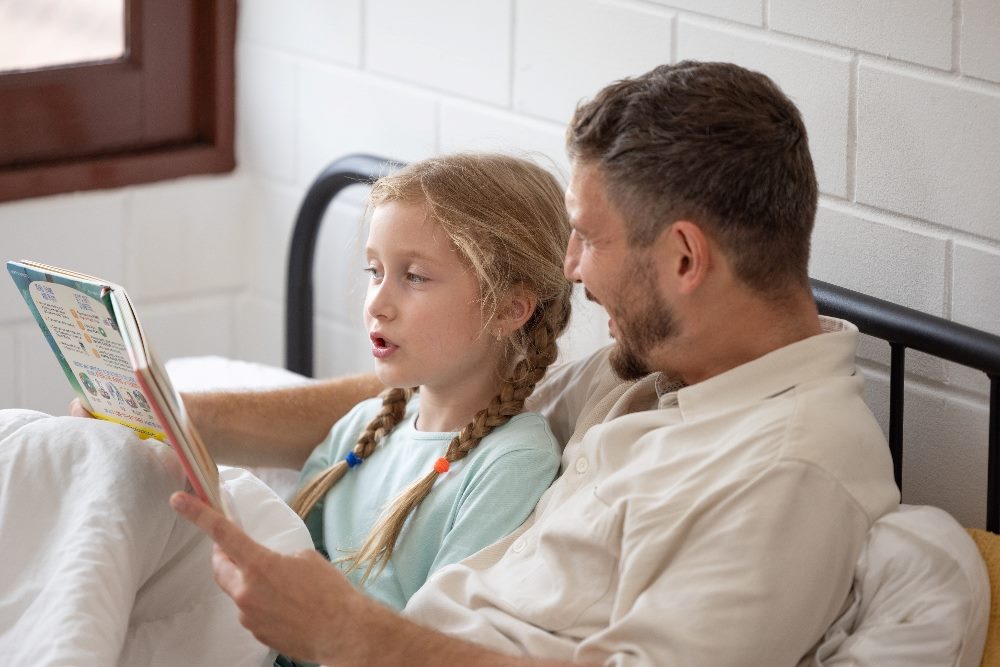 A father, who has a beard and is wearing a white shirt ,reads to his red-haired daughter, who is wearing her hair in two braids, from a storybook in piece about brand storytelling.