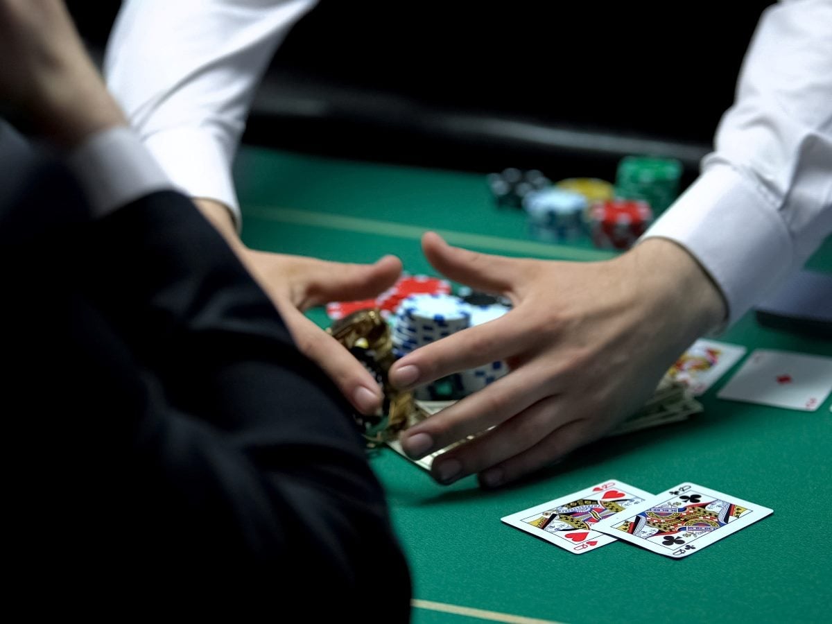 Close-up of a casino dealer’s hands collecting poker chips and cards across a green table while players sit nearby.
