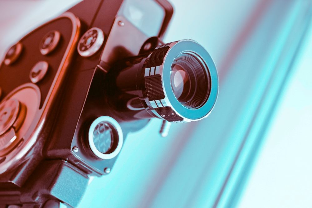 A close-up shot of a vintage film camera, highlighting the lens and dials. The image features a cool blue and pink color gradient, giving it a modern and artistic look. The background is softly blurred, drawing attention to the camera's intricate details in piece about video content.