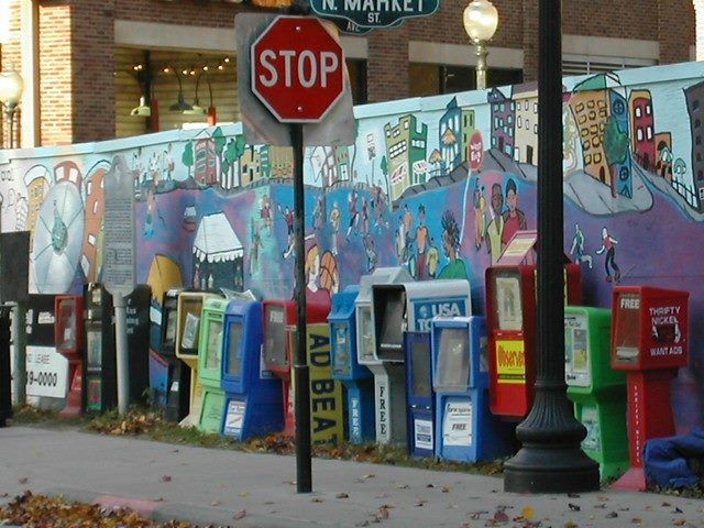 Row of newspaper boxes on a street behind a stop sign.