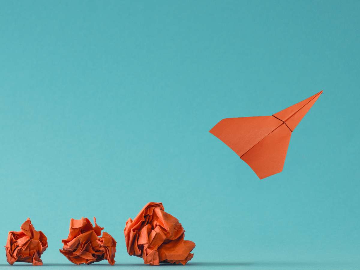 Four stages of transformation shown with orange paper: three crumpled balls on the ground and one paper airplane flying upward against a blue background, symbolizing progress and innovation.