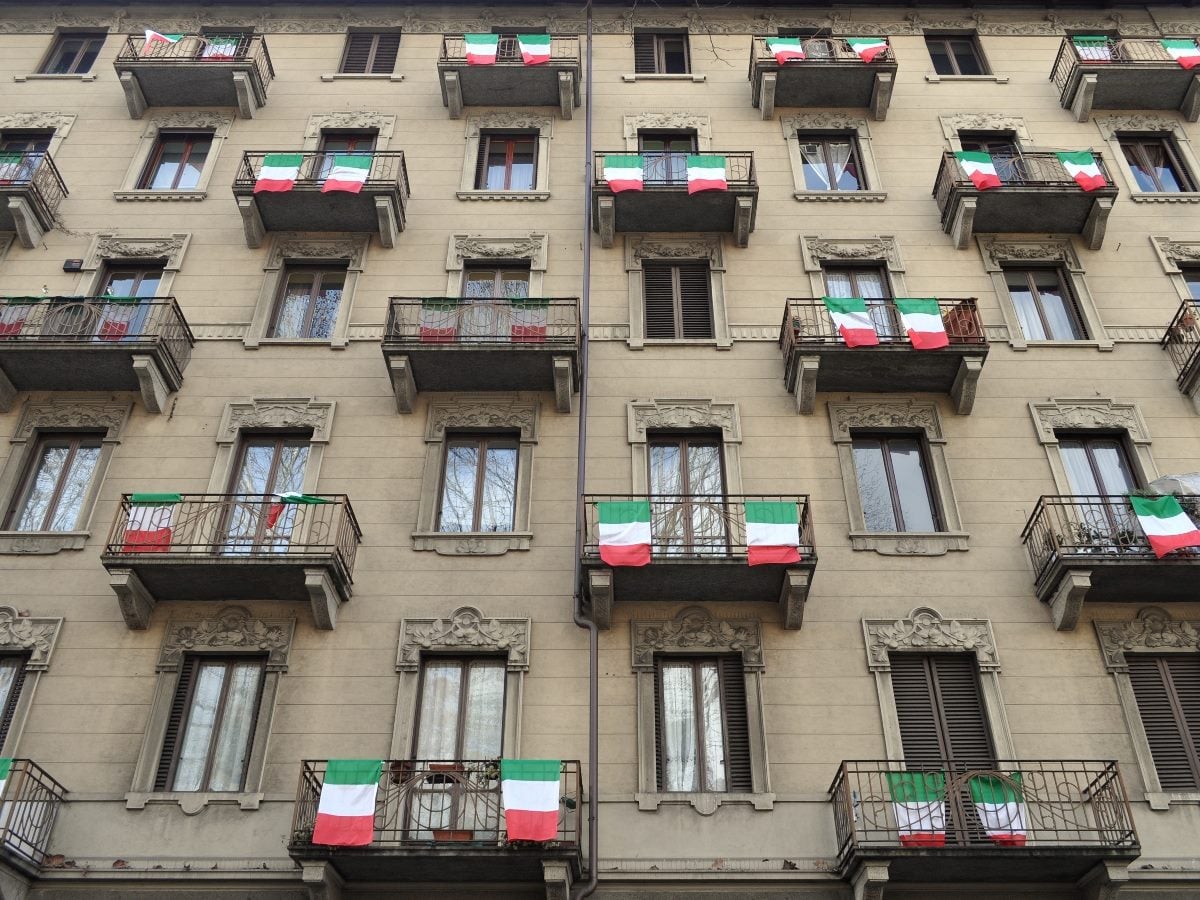 The straight-on facade of a classical European apartment building, five stories tall, built from beige stone with ornate carved window surrounds, floral relief details and decorative cornices. Wrought-iron balconies extend from windows across every floor, and Italian tricolor flags — green, white and red — hang draped over roughly a dozen of the balcony railings in a loosely repeating pattern. Tall shuttered windows with dark wood frames line the lower floors, while upper windows show sheer curtains and glimpses of leafless tree branches reflected in the glass. A vertical drainpipe runs down the center of the frame, dividing the facade into two roughly symmetrical halves. The photo is taken from street level at a slight upward angle, in flat, even daylight with sharp focus across the entire building and no visible sky or foreground.