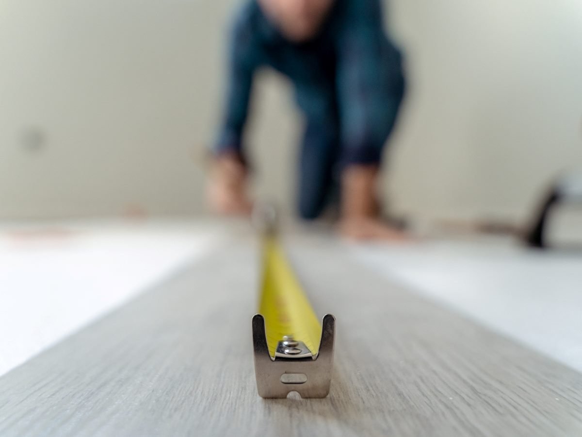 Close-up view of a measuring tape stretched across a floorboard, with a person in the background kneeling and working, emphasizing precision and evaluation.