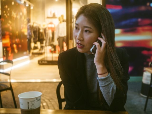 Woman talking on phone outdoors in front of store with a cup of coffee on the table in front of her. 