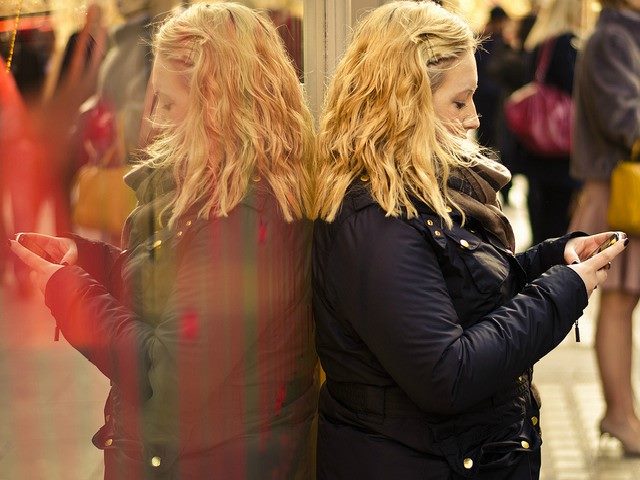 woman reading mobile phone leaning against a glass storefront with her reflection showing.