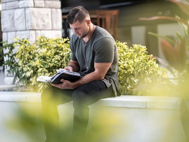 Former NFL quarterback Tim Tebow sitting down reading a book outside.