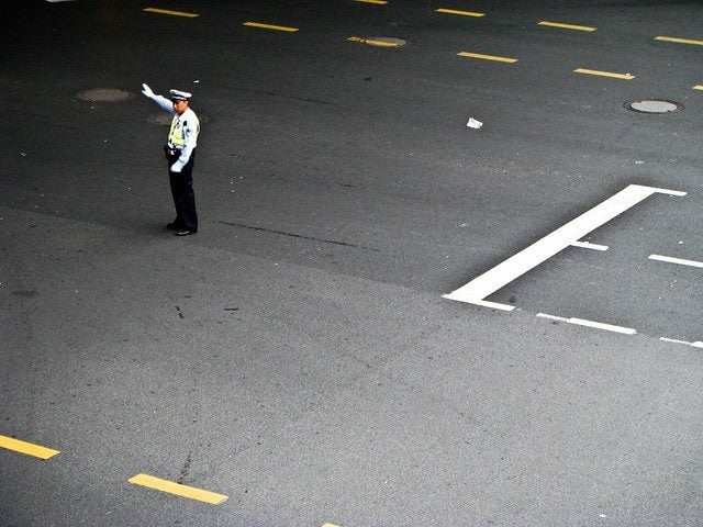 A traffic guard stands in the middle of the street with his one hand up, signalling someone to stop.