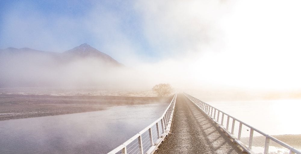 This image shows a bridge stretching across a foggy river landscape with a mountain in the background, bathed in soft sunlight. The bridge symbolizes connection and transition, much like customer advocacy efforts that bridge the gap between a brand and its consumers, guiding them through their journey with the brand.