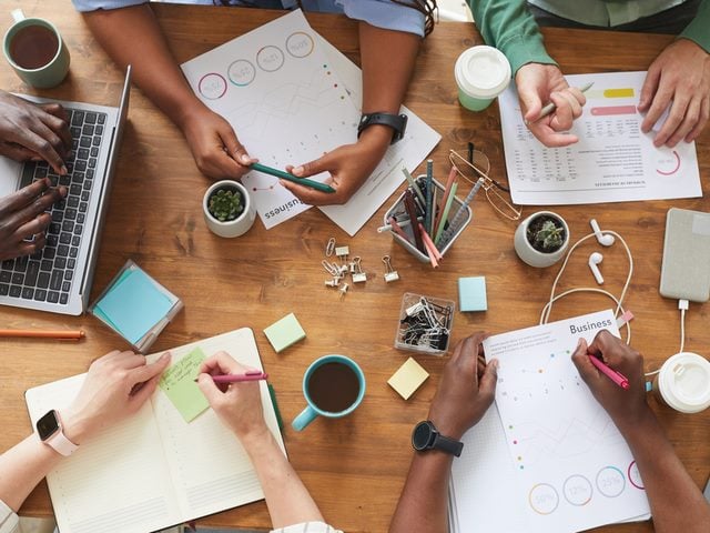 Top view close up of multi-ethnic group of people working together at cluttered wooden table with coffee cups, mugs and stationary items.