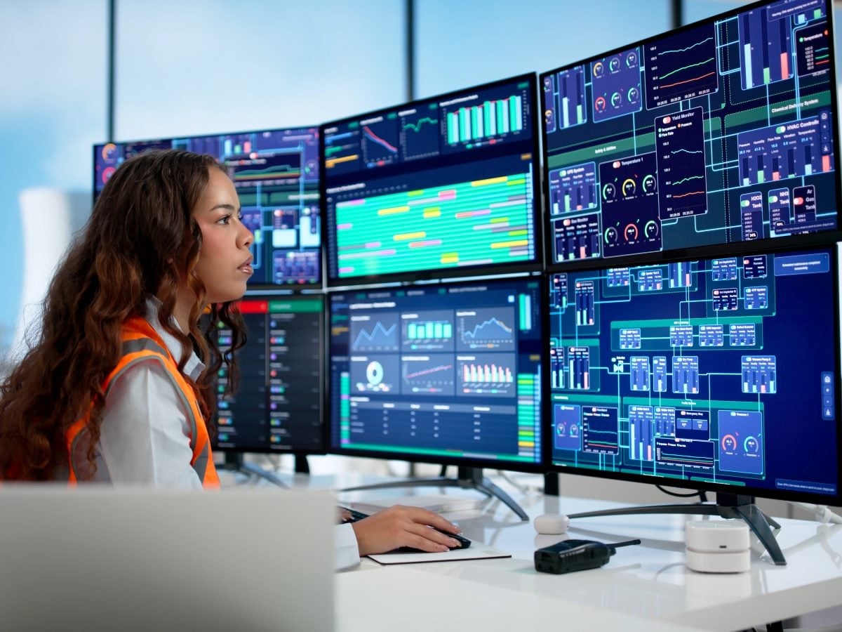 A woman with long curly brown hair, wearing an orange safety vest over a white top, sits in profile at a white desk in a bright control room, focused intently on a wall of six large monitors arranged in a grid displaying industrial monitoring dashboards with dark blue backgrounds and teal, orange, pink and green data visualizations, including line charts, bar graphs, circular gauges and system diagrams labeled with terms like Yield Monitor, HVAC Controller, Chemical Delivery System and Gas Monitor; her right hand rests on a computer mouse on the desk, which also holds a two-way radio and a small white charging dock, with a laptop visible in the foreground and a white office chair to her left; large windows behind her show a hazy blue sky and the blurred cooling towers of an industrial or power plant in the background, with soft, even daylight illuminating the scene and a shallow depth of field that keeps the woman and nearby screens sharp while the background towers and windows soften into a blur.