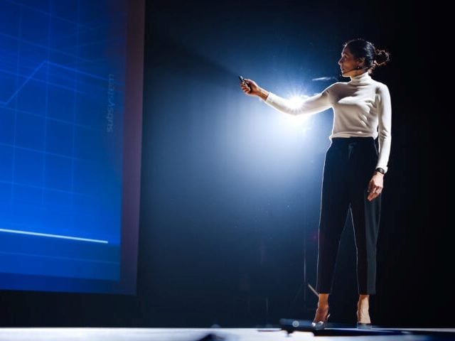 A woman stands giving a presentation at a Medallia Experience conference in piece about Medallia Experience '24.