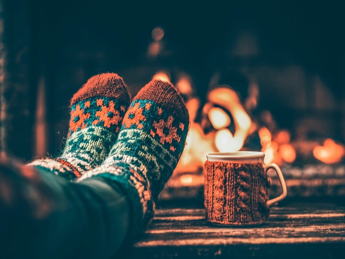A person with their feet up, wearing cozy socks with a coffee mug next to their feet and fireplace in the background, signaling a holiday season scene.