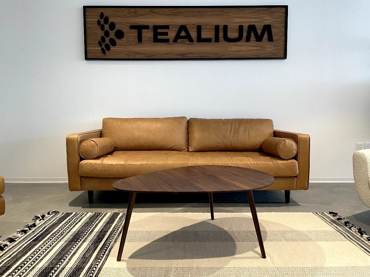 A modern office seating area with a tan leather sofa, round bolster pillows and a dark wood coffee table on a patterned rug. A wooden wall sign above the couch reads "Tealium" in bold black letters.