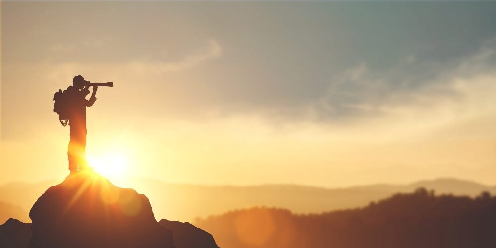 A person looks through a long monocular while standing on a rock with a dramatic sunset in the background in piece about Adobe Summit Insights 2024.