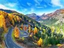 A winding mountain road curves through a valley filled with vibrant autumn foliage in shades of orange, yellow, and green surrounded by tall peaks under a bright blue sky.