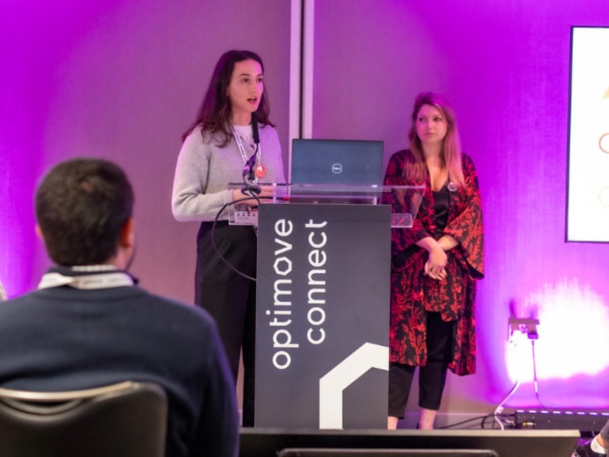Two women present on stage at an Optimove Connect event, with one speaking at a podium and the other standing beside her, addressing an audience under purple conference lighting.