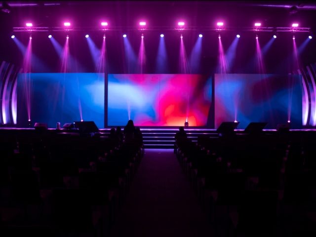 Front view of the stage event with a big LED screens and purple and pink lights are shining down on the stage floor while testing the light system with many empty chairs arranged for audience in hall in piece about contact center conferences and events. 