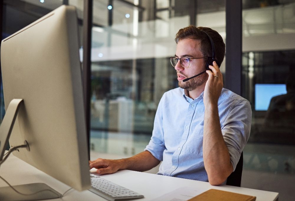 A customer service employee sits in front of a desktop computer and speaks while wearing headphones in a modern glass and steel office setting in piece about AI in customer experience and customer service.