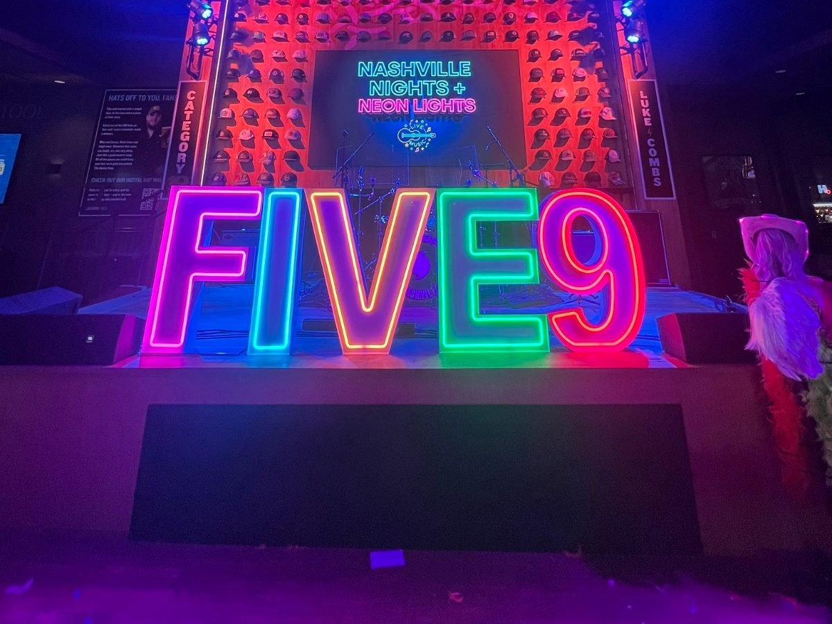 A large neon sign reading “FIVE9” glows in pink, blue, green and red in front of a stage at Five9’s CX Summit in Nashville. The backdrop features a wall of hats and a screen displaying “Nashville Nights + Neon Lights.” Stage lights cast colorful reflections across the venue, where a scarecrow type of figure dressed in bright, feathered accessories stands to the right.