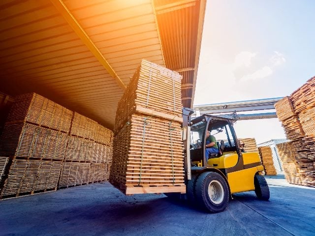 Forklift loader with a load of lumber surrounded by wood in drying in containers. 