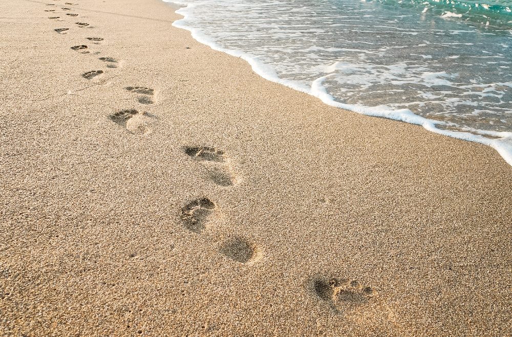 The image shows a sandy beach with a line of footprints leading toward the horizon. The foamy edge of a wave gently washes onto the shore, partially covering the lowest footprints. The scene captures a tranquil, natural setting under a bright, clear sky in piece about the impact of micro-influencers on Gen Z for Amazon Prime Day.