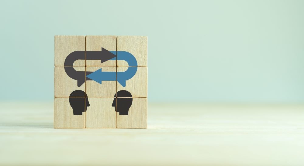 Wooden blocks stacked together form an image of two people's heads in conversation wit a black and blue loops with two arrows above them in piece about employee and customer feedback loops.